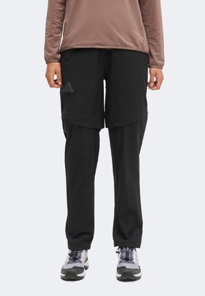Personne portant un pantalon convertible noir avec short zippé, un haut marron à manches longues et des chaussures de sport grises, debout devant un fond uni.