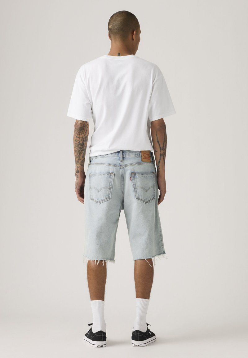 Man met tatoeages draagt een wit t-shirt, lichtblauwe versleten spijkershorts, witte sokken en zwarte sneakers, en staat met de rug naar de kijker op een effen achtergrond.