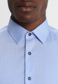 Camicia azzurro chiaro con texture liscia, colletto classico e bottoni scuri. Presenta un motivo sottile nel tessuto e una vestibilità su misura.