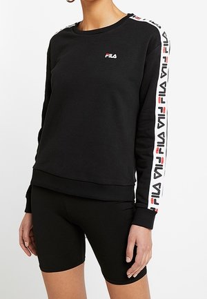 Femme portant un sweat-shirt FILA noir avec des bandes blanches marquées sur les manches et un short noir ajusté, debout devant un fond uni.