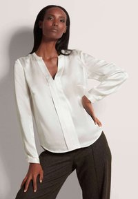 Weißseide Bluse mit V-Ausschnitt, langen Ärmeln und subtiler Glanz. Der Stoff fällt sanft und ergänzt maßgeschneiderte dunkle Hosen.