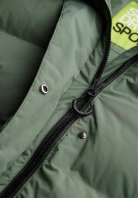 Grüne Puffjacke mit schwarzem Reißverschluss und Beschlägen. Verfügt über einen strukturierten Stoff, einen runden Logo-Reißverschlusszieher und ein leuchtend gelbes Etikett innen.