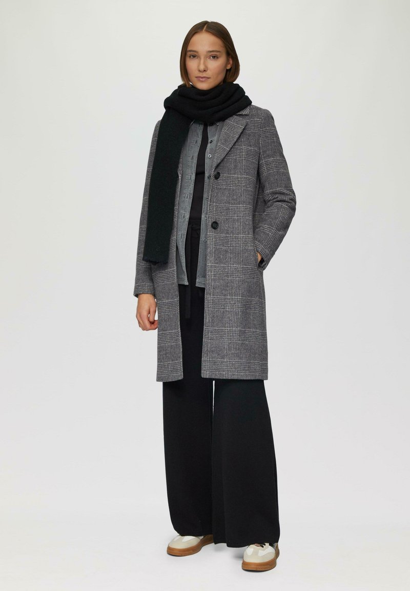 Manteau à carreaux gris avec des boutons double boutonnage, associé à une écharpe noire, un haut noir, un pantalon large et des baskets beiges.