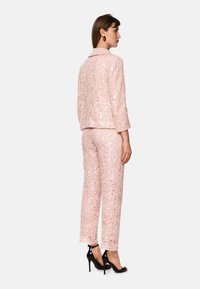 Helles rosa Paillettenset mit einem taillierten Blazer und geraden Hosen, versehen mit einem strukturierten Spitzenmuster und zwei vorderen Taschen, kombiniert mit schwarzen Absatzschuhen.