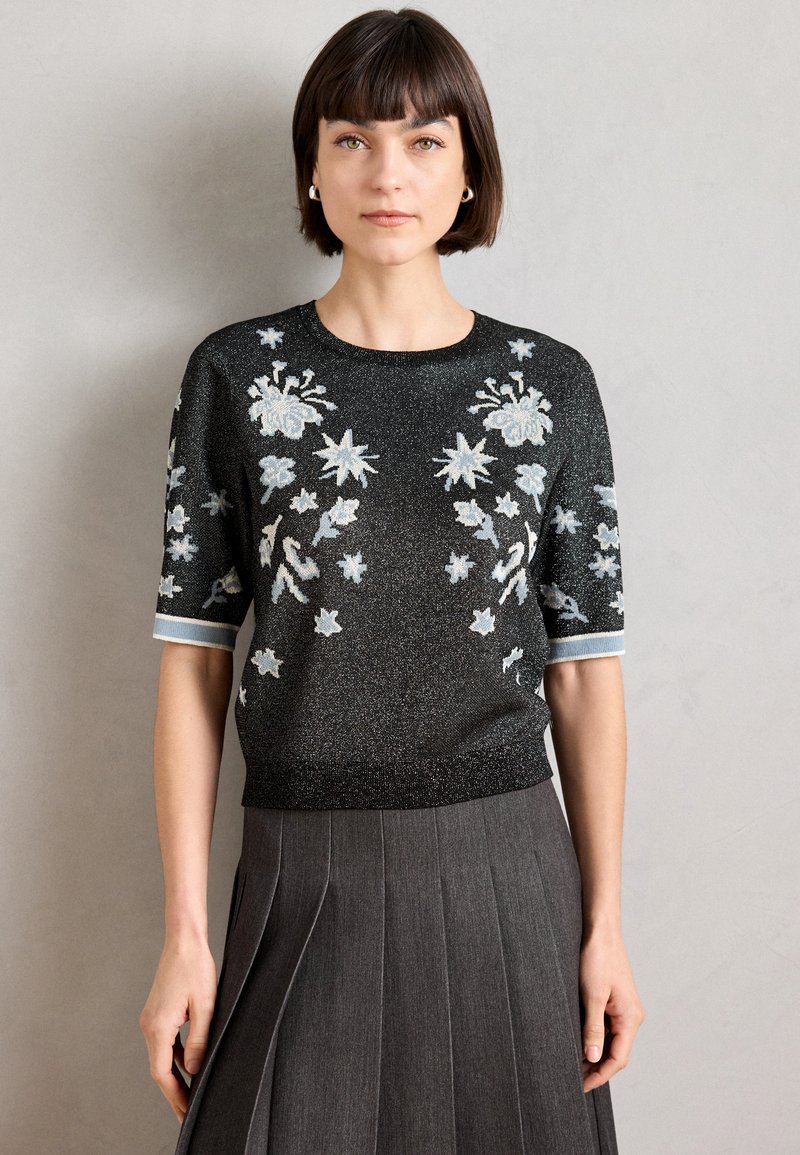 Μαύρη κοντομάνικη μπλούζα με λουλουδάτη κεντητική σε ανοιχτό μπλε και ασημί, διαθέτει στρογγυλή λαιμόκοψη και ραμμένα μανίκια, συνδυασμένη με μια πλισέ γκρίζα φούστα.