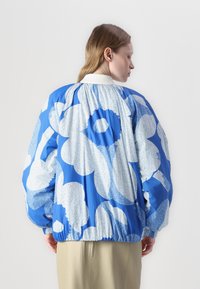 Casaco azul oversized com grandes padrões florais brancos. Apresenta um costas franzida e punhos elásticos. Tecido liso e leve.