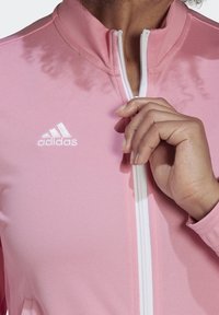 Chaqueta adidas rosa con cremallera frontal, cuello alto y puños acanalados. Presenta una textura en relieve y un pequeño logo blanco en el lado izquierdo del pecho.
