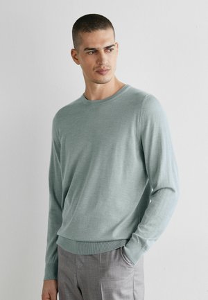 Mężczyzna ubrany w jasnooliwkowy sweter z długim rękawem i szare spodnie, stoi z jedną ręką w kieszeni, patrząc w lewo na tle białej ściany.