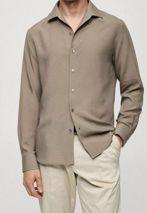 Homme portant une chemise taupe à manches longues boutonnée avec un pantalon beige, une main dans la poche et l'autre pendant.