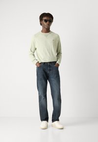 Pulover lung cu mâneci lungi de culoare verde deschis, cu tricot texturat, asortat cu blugi drepti de culoare bleumarin și pantofi albi; ochelarii de soare completează look-ul.