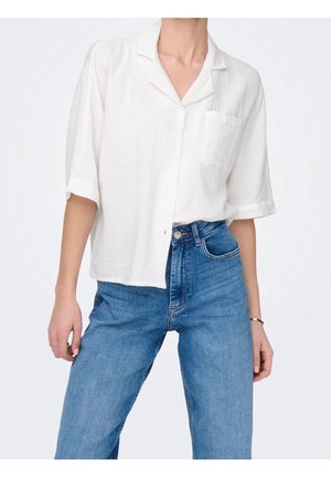 Vrouw draagt een losse witte korte-mouwen blouse met knopen, ingestopt in een blauwe high-waisted spijkerbroek met een armband om de rechterpols.