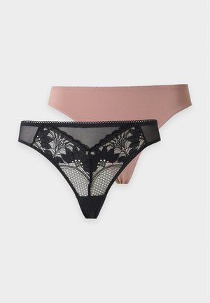 Mujer de pie descalza con un sujetador beige y bragas malva sobre un fondo liso gris claro.