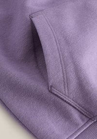 Gros plan sur une poche de sweat à capuche lavande avec des coutures fines et un poignet côtelé visible sur le bord.