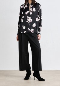 Blouse noire à imprimé floral avec manches longues et col noué, associée à un pantalon noir large et des bottines noires pointues.