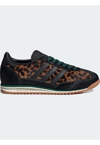 Baskets basses avec imprimé léopard et accents en cuir noir, lacets verts, semelle beige et trois bandes Adidas sur le côté.