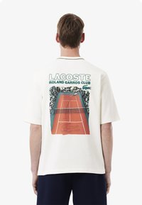 T-shirt bianca a maniche corte con una grafica retro di una scena di tennis, con testo "LACOSTE ROLAND GARROS CLUB" e dettagli verdi.