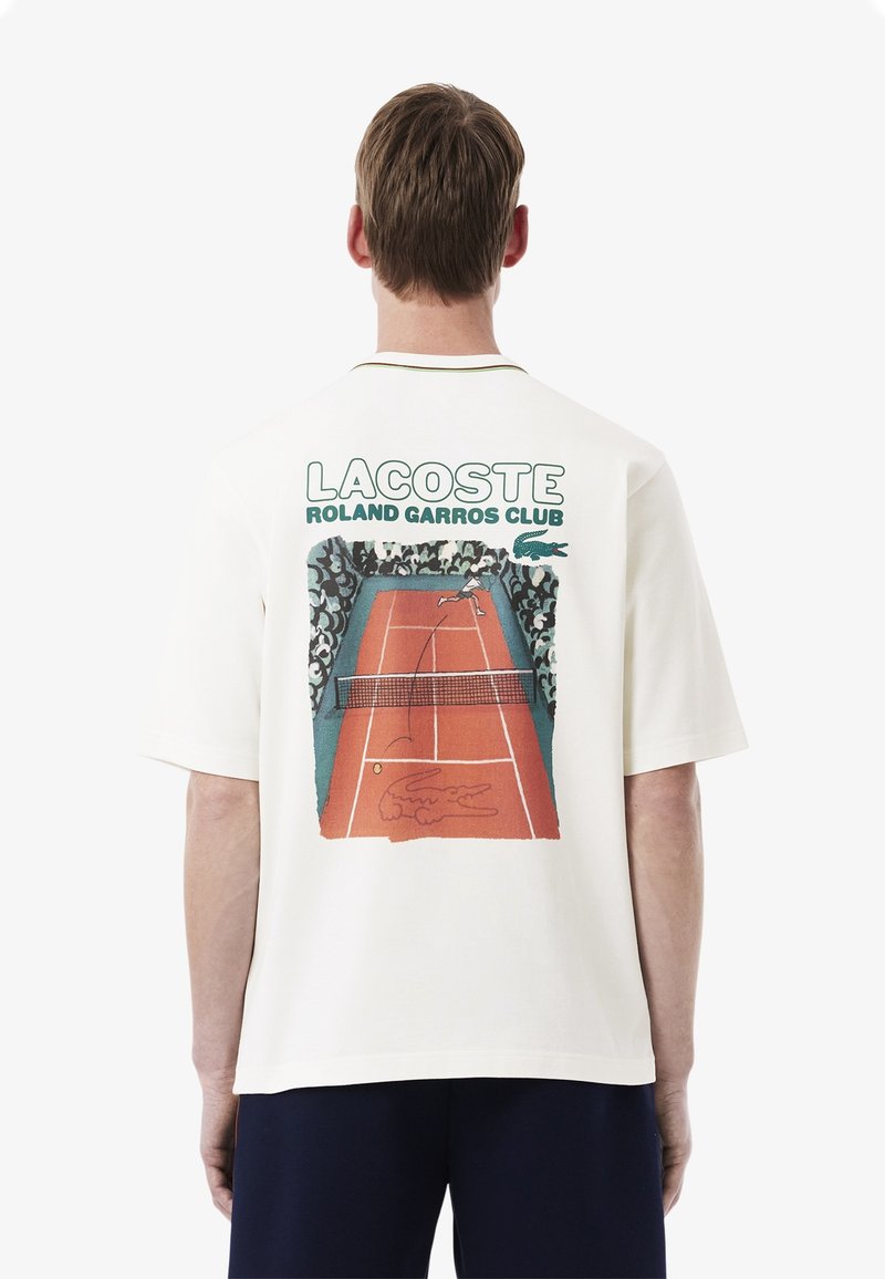 T-shirt bianca a maniche corte con una grafica retro di una scena di tennis, con testo "LACOSTE ROLAND GARROS CLUB" e dettagli verdi.