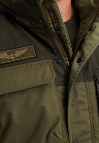 Grön militärjacka i stil med dragkedja, bröstficka med emblem och flera fickor med en texturerad tygfinish.