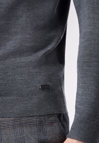 Donkergrijze gebreide trui met geribbelde manchetten en onderkant, met een klein zwart logo-label. Daaronder zijn de zichtbaar geruite broeken.