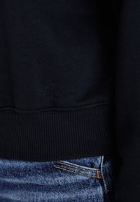 Close-up da bainha de um sweatshirt preto com um discreto logótipo bordado acima do punho canelado, combinado com jeans de ganga azuis com costura laranja.