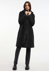 Zwarte fleece jas met een capuchon, voorzien van knopen aan de voorkant, twee zijzakken en een knielengte ontwerp. Gecombineerd met donkergrijze broek en laarzen.