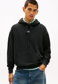 Felpa nera con cappuccio con tasca a marsupio, coulisse e una piccola toppa con logo, abbinata a una camicia verde chiaro con colletto sotto.