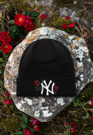 Berretto nero lavorato a maglia con logo bianco NY e ricamo di rosa rossa, posato su una roccia coperta di licheni circondata da petali di rosa rossi sparsi e vegetazione.