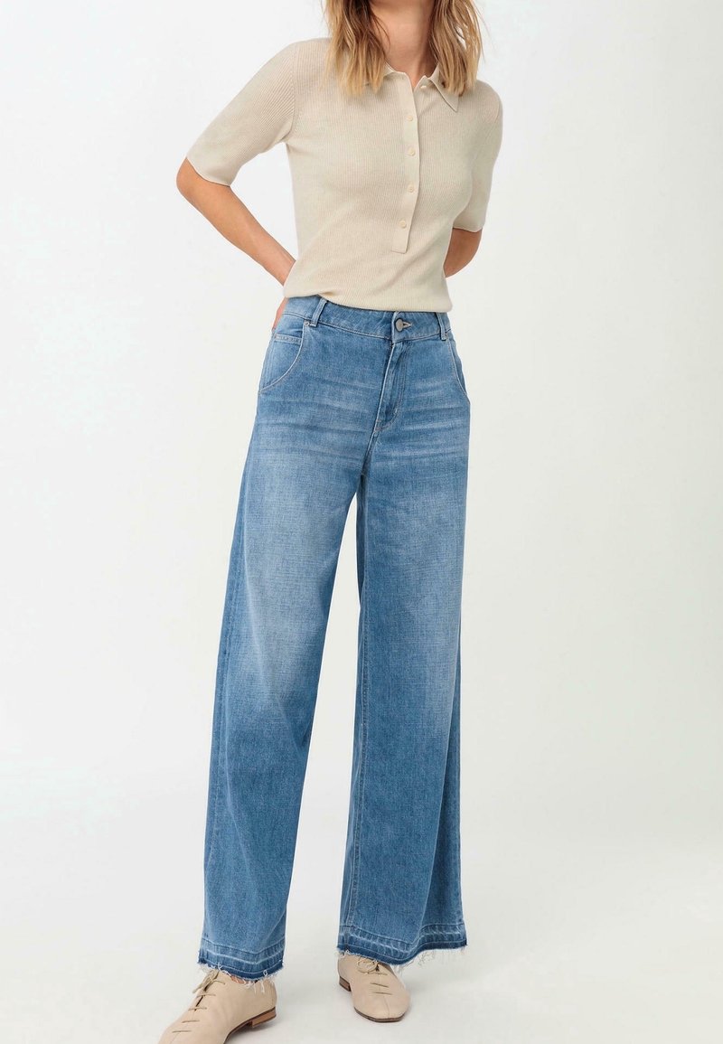 Polo en beige clair à manches courtes avec une patte de boutonnage, associé à un jean en denim bleu à taille haute et jambes larges, avec un ourlet effiloché.