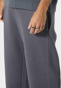 Pantalon de survêtement gris avec une texture lisse, featuring des poches latérales et des coutures contrastantes. Une main avec des bagues est placée près de la taille.