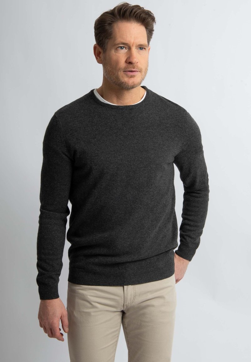 Donkergrijze wollen crewneck trui met ribgebreide mouwen en onderrand, met een klassiek ontwerp. Gedragen over een wit shirt, gecombineerd met lichtgekleurde broek.