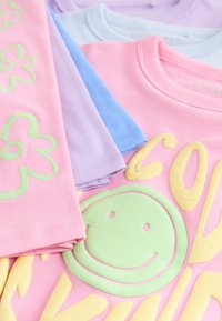 Pile de t-shirts de couleur pastel avec des imprimés de fleurs vertes sur des manches roses et un grand visage souriant vert avec du texte jaune sur un t-shirt rose.