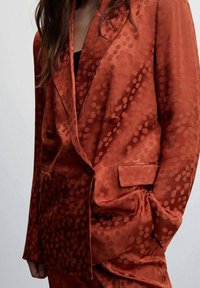 Femme portant un blazer en satin couleur rouille avec un motif à pois subtil, une main dans la poche, et de longs cheveux bruns visibles.