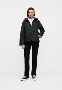 Γυναίκα που στέκεται, φοράει μαύρο καπιτονέ μπουφάν, λευκό πουκάμισο, μαύρο παντελόνι και μαύρα αθλητικά παπούτσια με λευκές σόλες και κορδόνια.