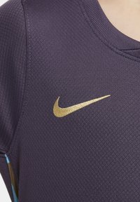 Maglietta sportiva Nike in tessuto viola scuro con una finitura testurizzata. Presenta un logo ricamato in oro sulla parte sinistra del petto e un colletto rotondo.