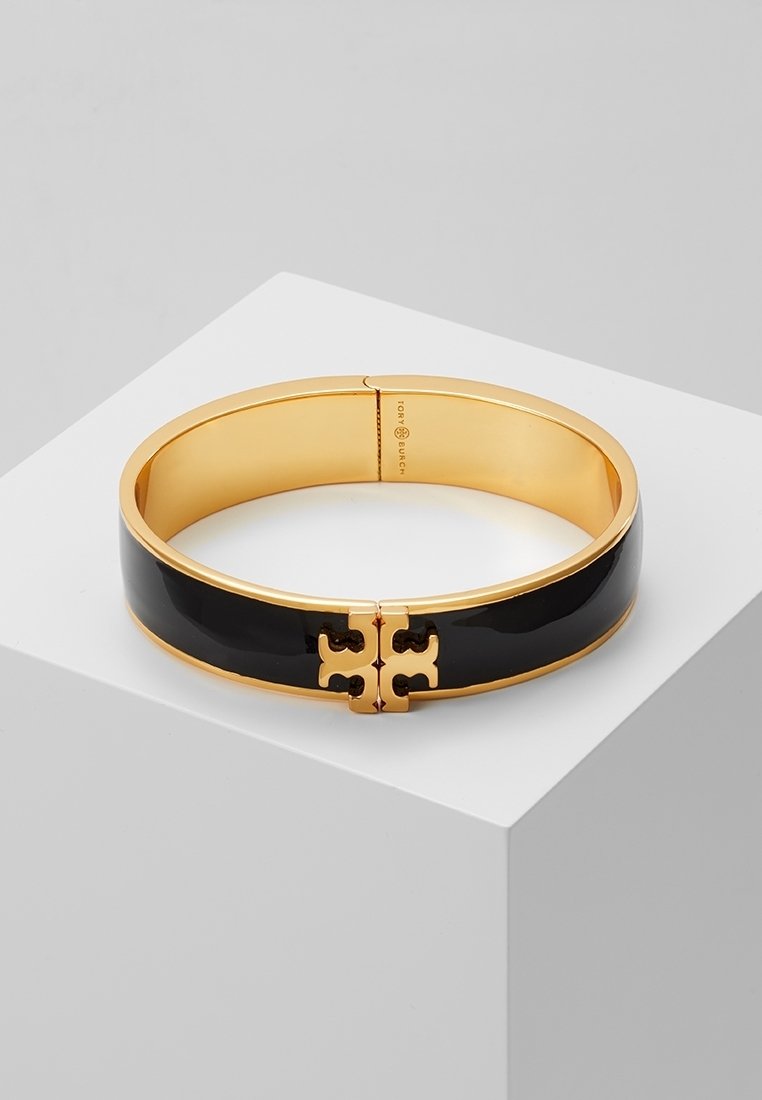 Bracelet jonc en émail noir et or avec fermoir logo doré entrelacé reposant sur une surface blanche.