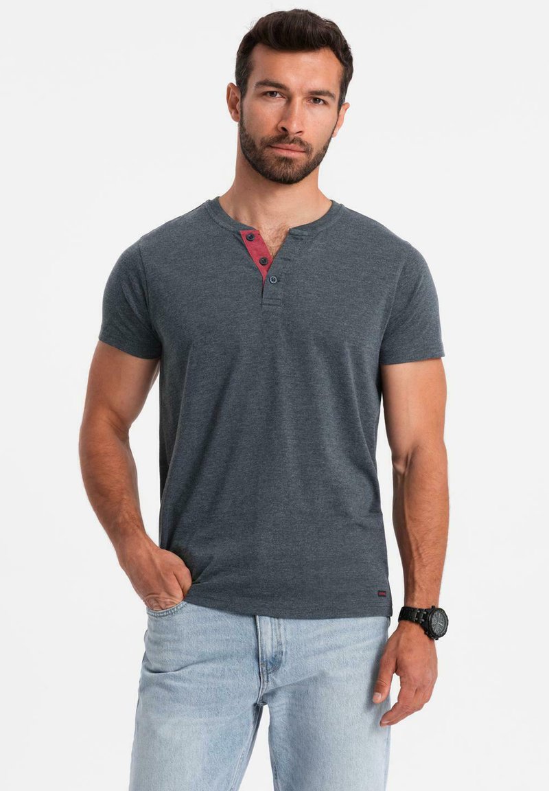 Mörkgrå Herr Henley-tröja med kort ärm som har en röd knappslå och två knappar. Bärs med ljusblå jeans, tyget verkar mjukt.