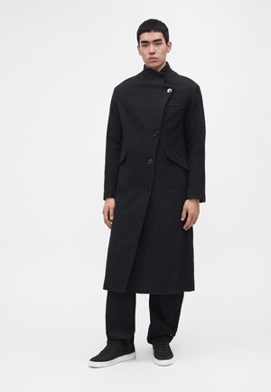 Jeune homme debout portant un long manteau noir asymétrique boutonné, un pantalon noir et des baskets noires à semelles blanches.