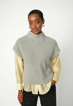 Top senza maniche grigio a maglia con un colletto alto, indossato sopra una camicia gialla chiara con bottoni. Dettaglio a coste testurizzato, vestibilità rilassata.