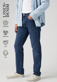 Hombre con jeans de mezclilla azul oscuro, camisa de mezclilla azul clara, zapatillas blancas, de pie con una mano en el bolsillo sobre un fondo gris.