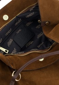 Braune Wildleder-Handtasche mit einem goldenen Reißverschluss und Druckknopfverschluss. Das Innere hat ein schwarzes Futter mit dem wiederholten "Venezia"-Logo und einem Lederschild. Glatte Textur.