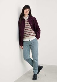 Burgundijas krāsas quiltota jaka virs krēma rakstainā džempera, gaiši zilas bikses un melni zābaki. Modelis balstās pret sienu neitrālā vidē.