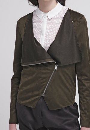Femme portant une veste zippée asymétrique vert olive sur un chemisier blanc à col avec un motif texturé.