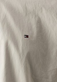 Lichtgrijs katoenen T-shirt met een klein geborduurd logo in rood, wit en zwart op de linkerborst, met een gladde textuur.