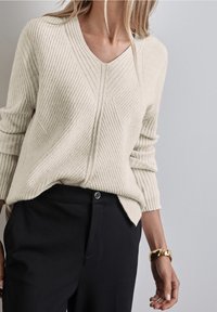 Pull crème en maille côtelée avec col en V et motif chevron texturé, associé à un pantalon noir avec fermeture à boutons.
