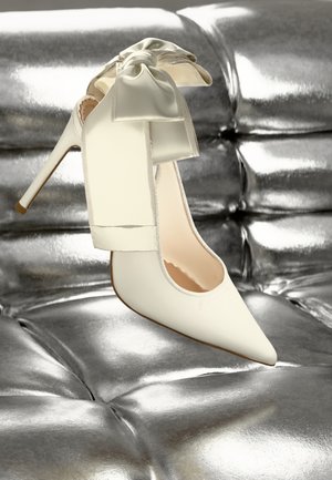 Talons aiguilles blancs en cuir lisse, ornés d'un grand nœud en satin à l'arrière, sur un fond métallique brillant.
