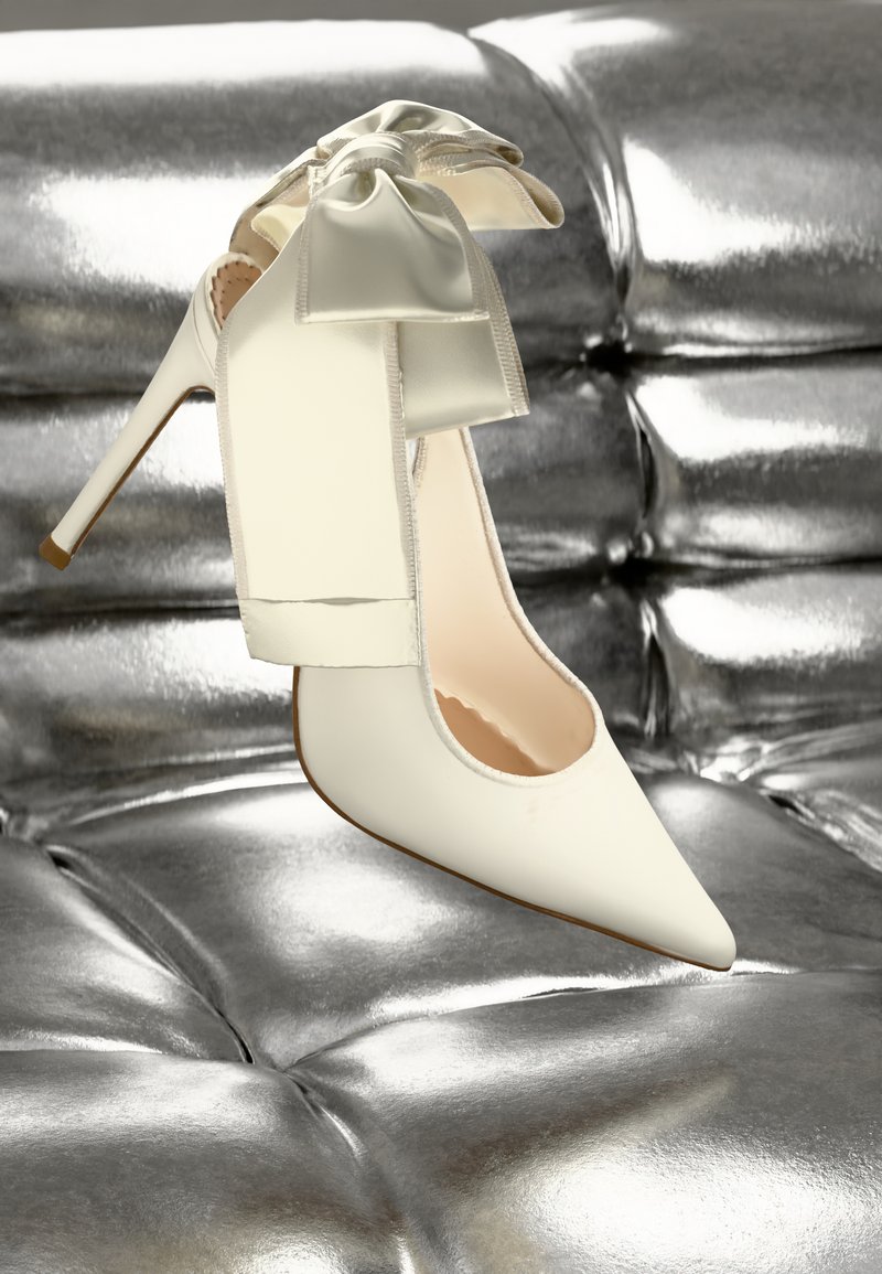 Talons aiguilles blancs en cuir lisse, ornés d'un grand nœud en satin à l'arrière, sur un fond métallique brillant.