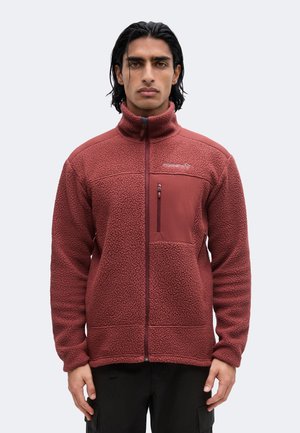 Hombre con chaqueta polar granate con cremallera completa, bolsillo con cremallera en el pecho y cuello alto, de pie frente a un fondo liso y claro.