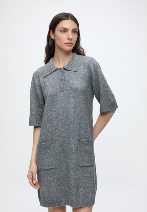 Femme aux longs cheveux foncés portant une robe en laine grise avec manches courtes, un col, trois boutons et deux poches avant.