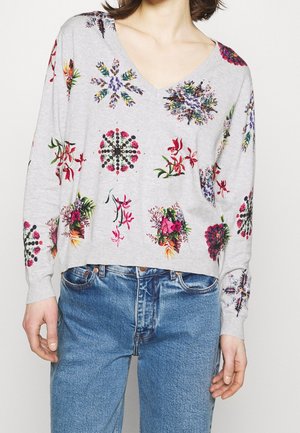 Femme portant un pull léger gris clair à col en V avec des motifs floraux multicolores, associé à un jean bleu, vue du cou jusqu'aux hanches.