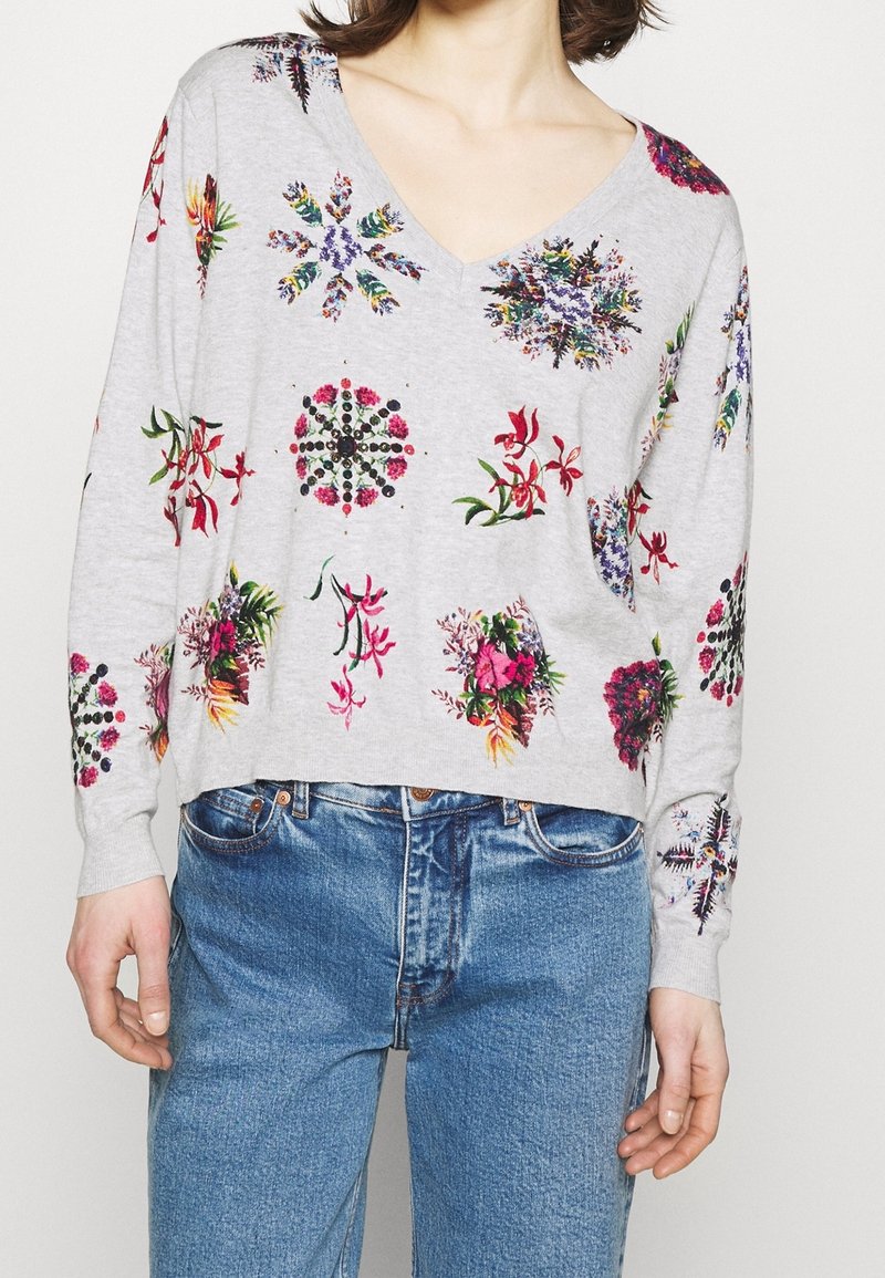 Femme portant un pull léger gris clair à col en V avec des motifs floraux multicolores, associé à un jean bleu, vue du cou jusqu'aux hanches.